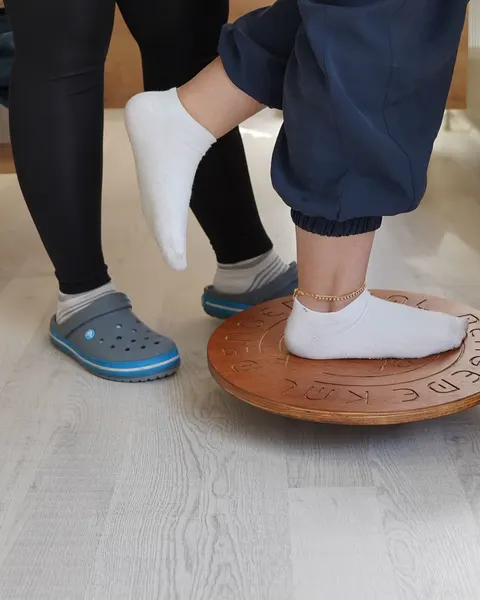 Füße auf einem hölzernen Balancekreisel, daneben die Therapeutin